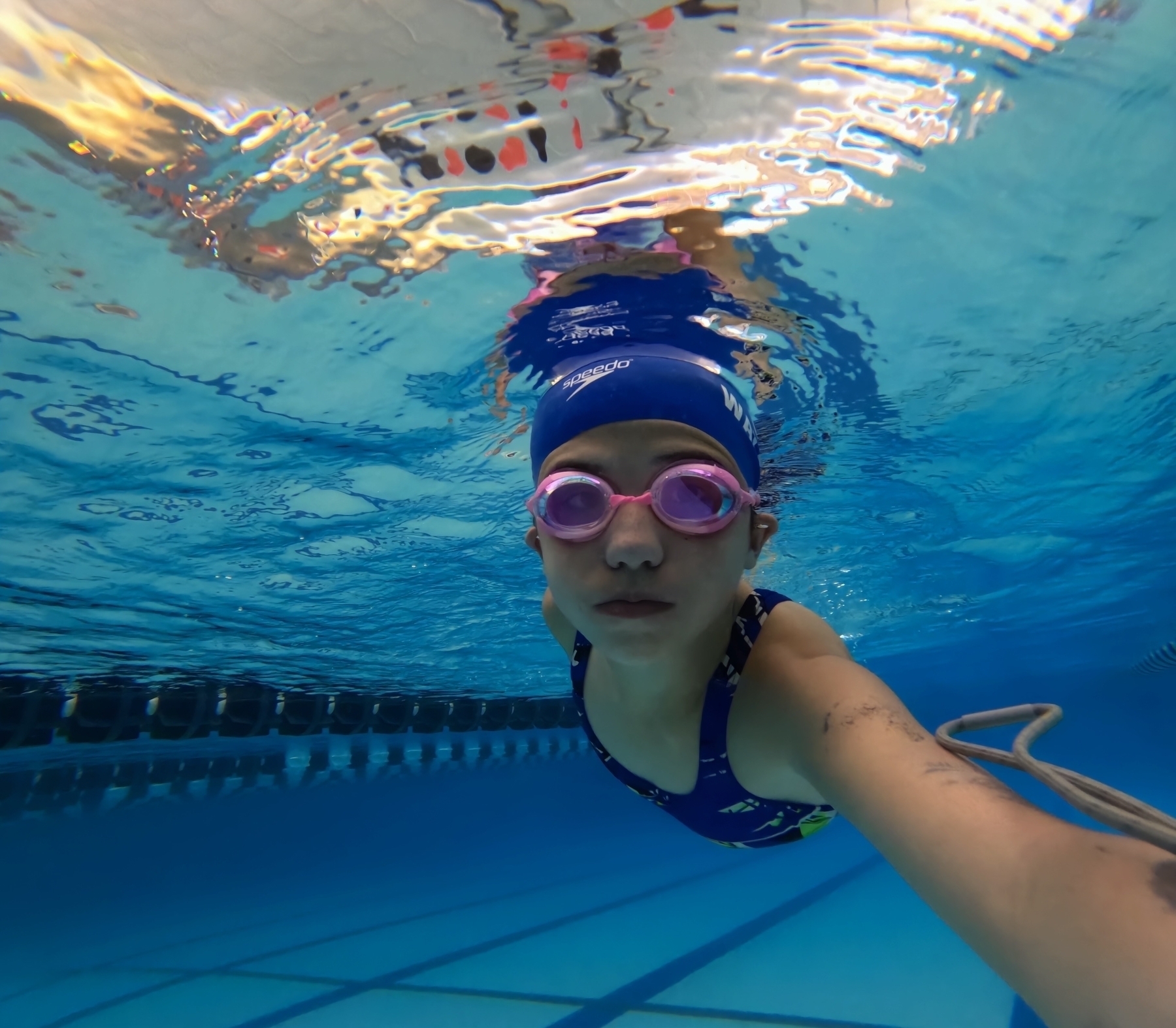 Person swimming underwater wearing pink goggles and a blue swim cap, taking an underwater selfie in a clear blue pool with lane lines visible.