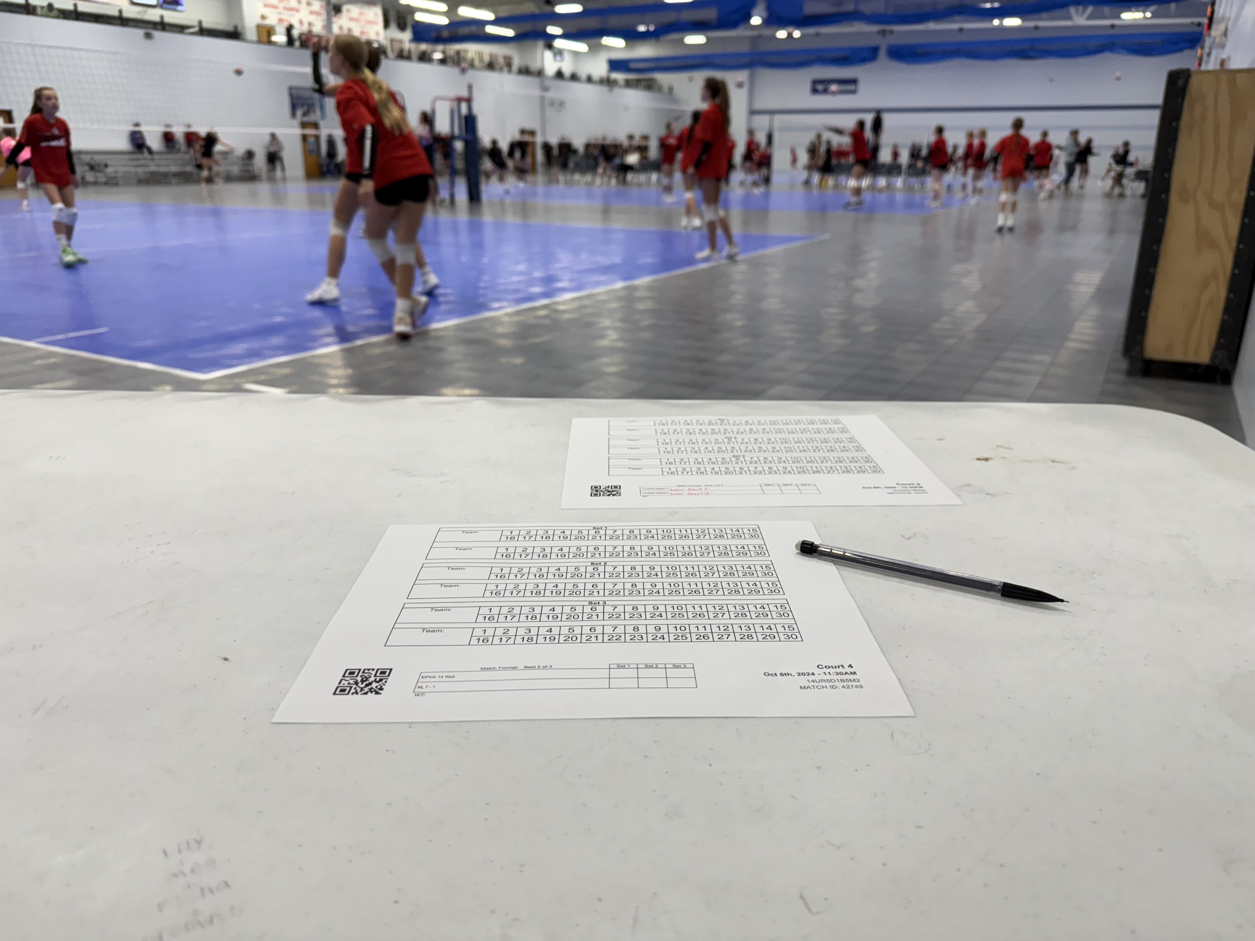 A volleyball court hosts players in red uniforms practicing, while a white table in the foreground holds tournament score sheets and a pen. The venue is spacious with multiple courts.