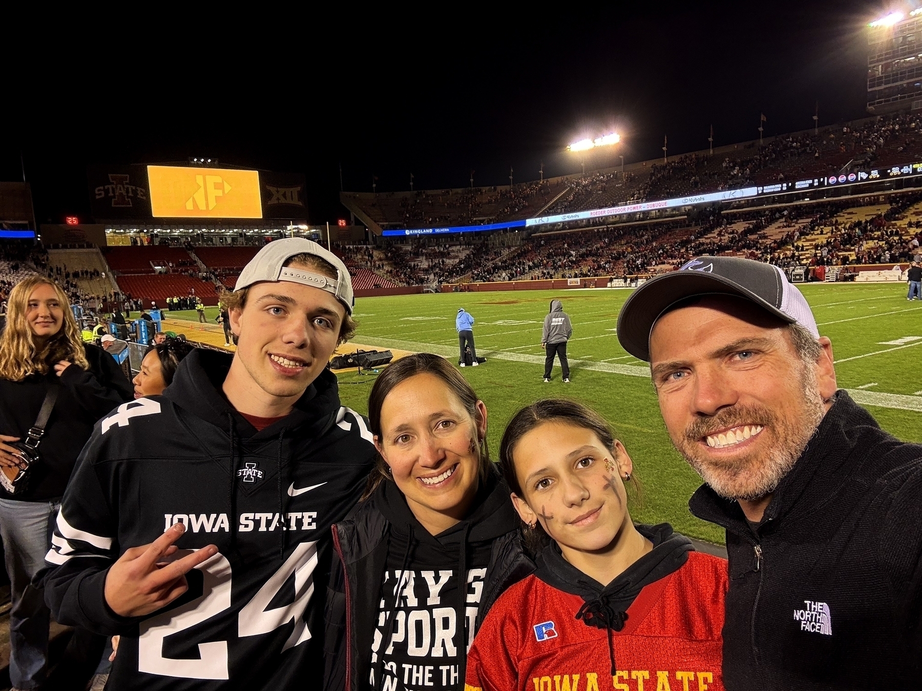 Four people smile for a group photo on a football field, surrounded by a stadium with spectators. One person wears an “Iowa State” jersey. The scoreboard and stadium lights are visible.