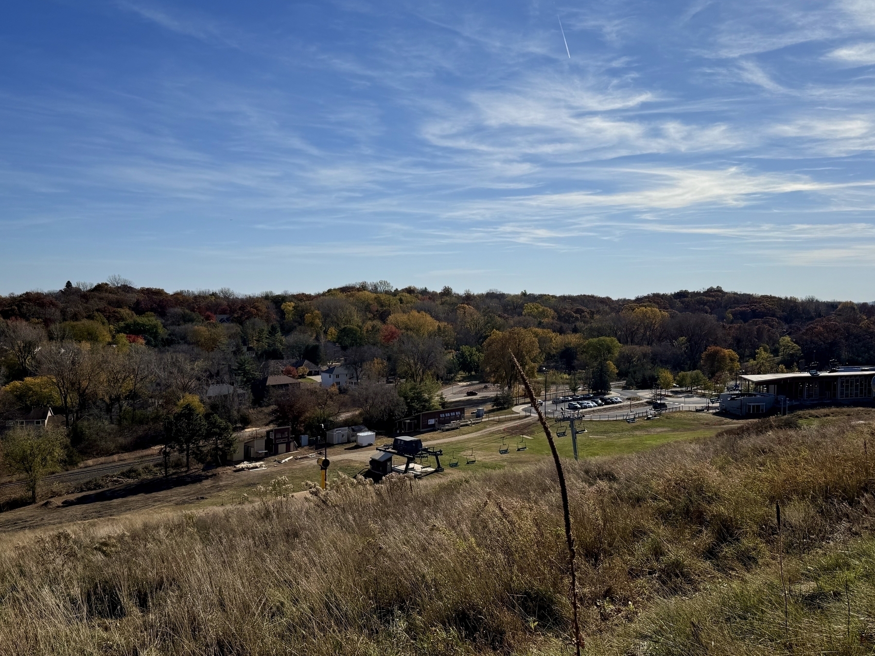 Grassy hillside overlooks a valley with scattered trees, buildings, and a parking lot. Ski lift chairs are stationary, indicating a recreational area. Blue sky with light clouds provides a serene backdrop.