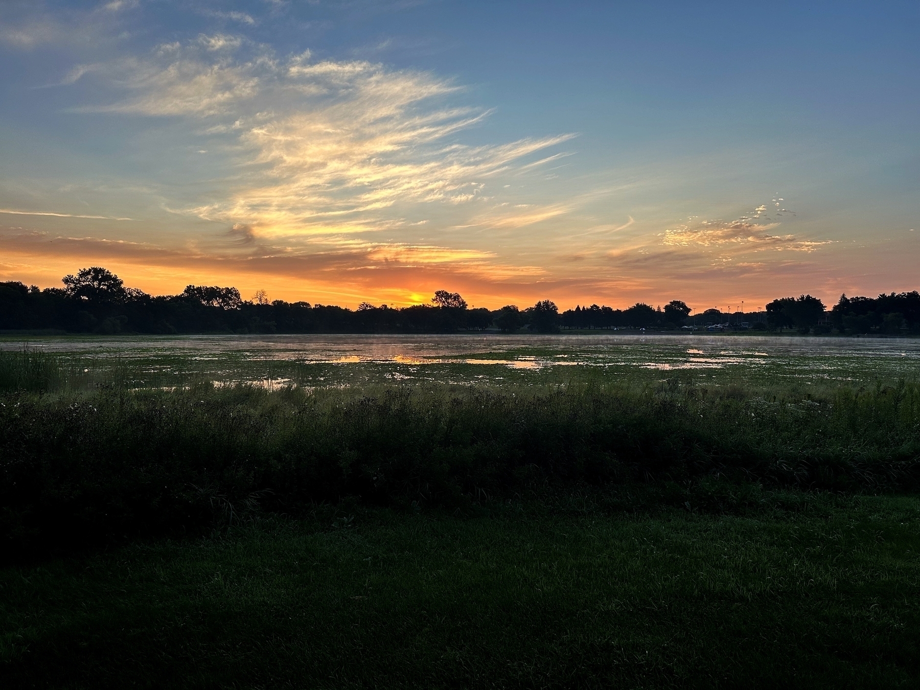 Sun sets over a tranquil lake, with orange and yellow hues lighting up the sky, surrounded by silhouetted trees and grassy fields reflecting the colorful twilight.