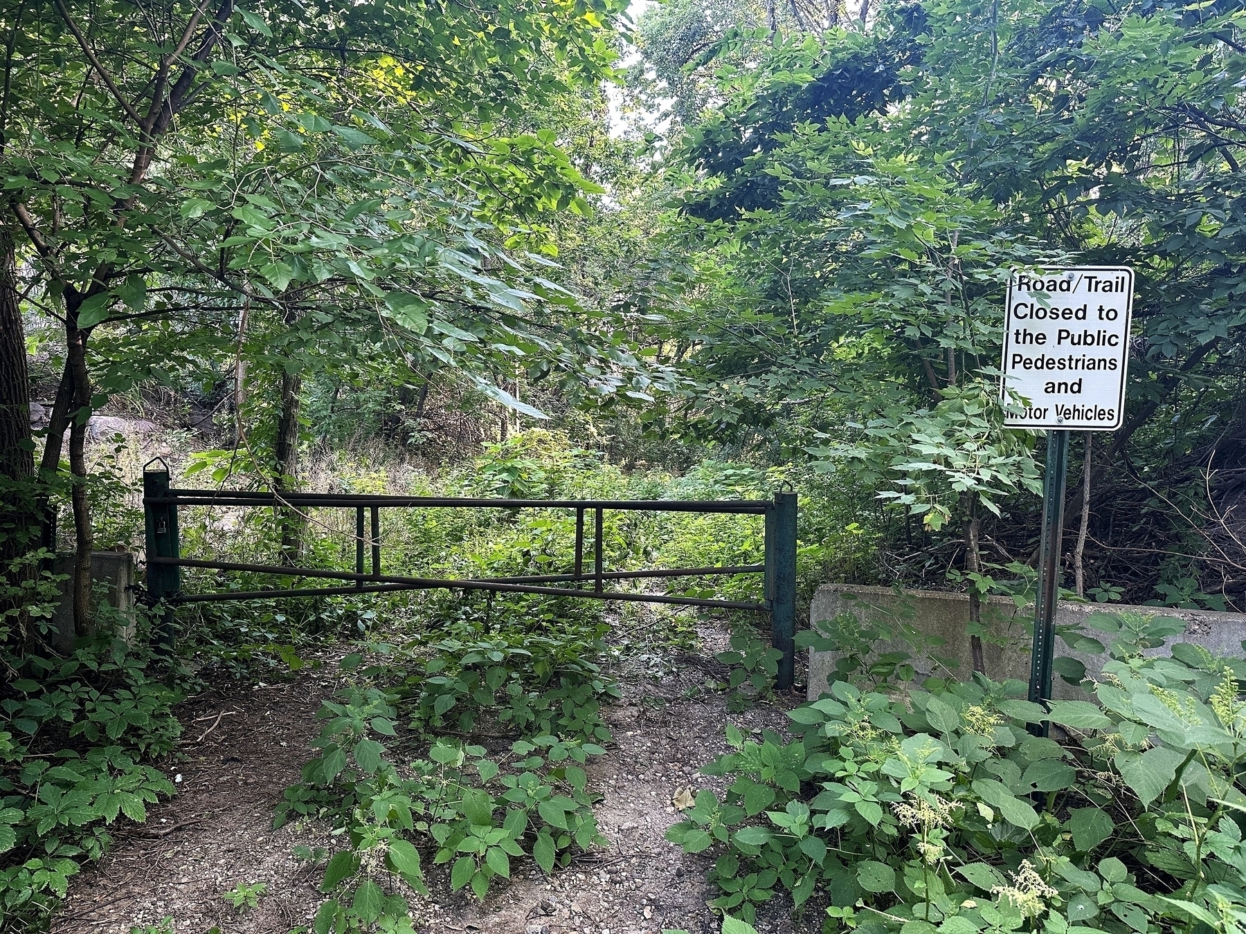 A metal gate blocks a dirt path surrounded by dense, green forest. A sign reads, “Road/Trail Closed to the Public Pedestrians and Motor Vehicles.