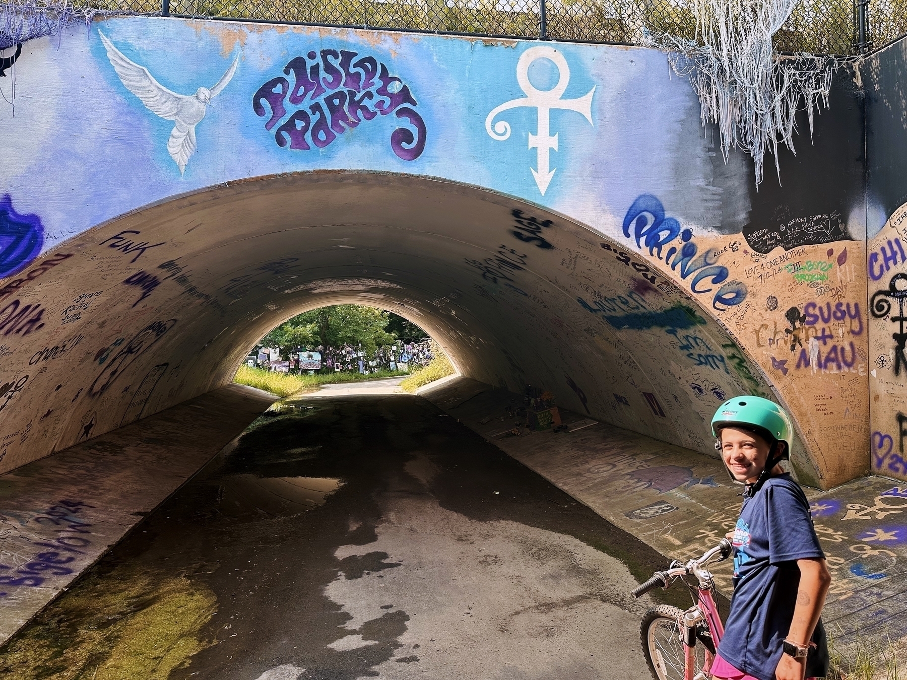 A child wearing a helmet stands with a bicycle in front of a graffiti-covered tunnel entrance with the words “Paisley Park” and “Prince”. A green, wooded area is visible through the tunnel.