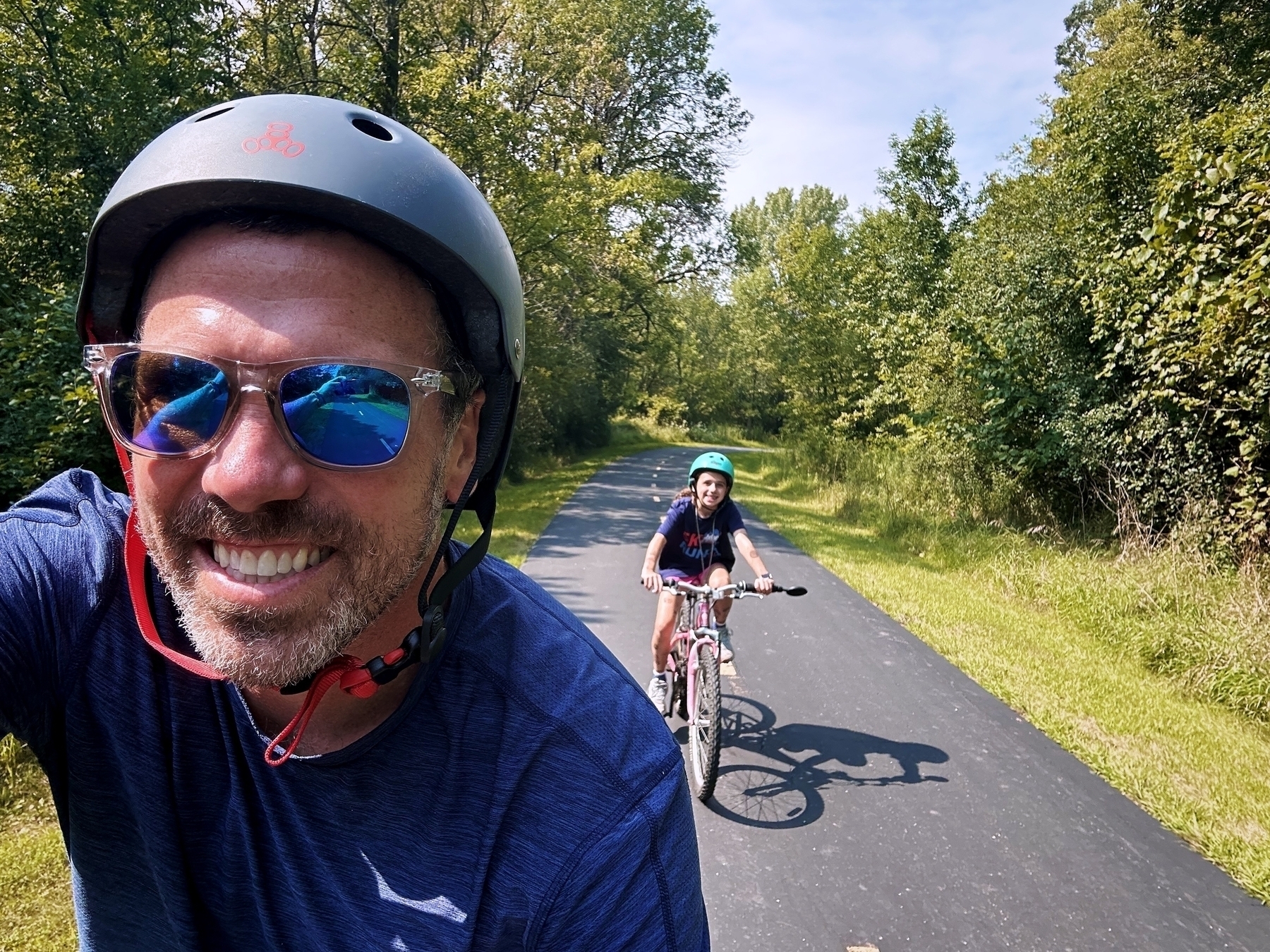 A man wearing sunglasses and a helmet smiles while biking on a paved path in a green, tree-lined park, with a young girl biking behind him.