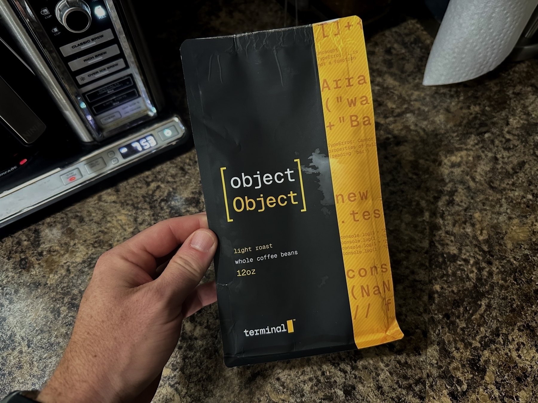 A hand holds a black and yellow coffee bag labeled “Object” with “light roast whole coffee beans 12oz” in a kitchen with a coffee maker on a granite countertop.