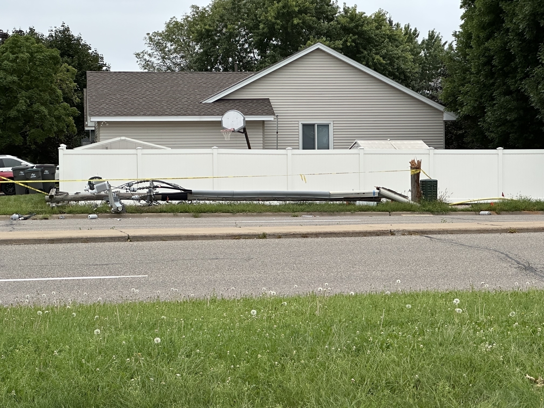 A fallen streetlight lies on a grassy roadside near a beige house with a white fence. Caution tape surrounds the scene, indicating recent damage or construction activity.