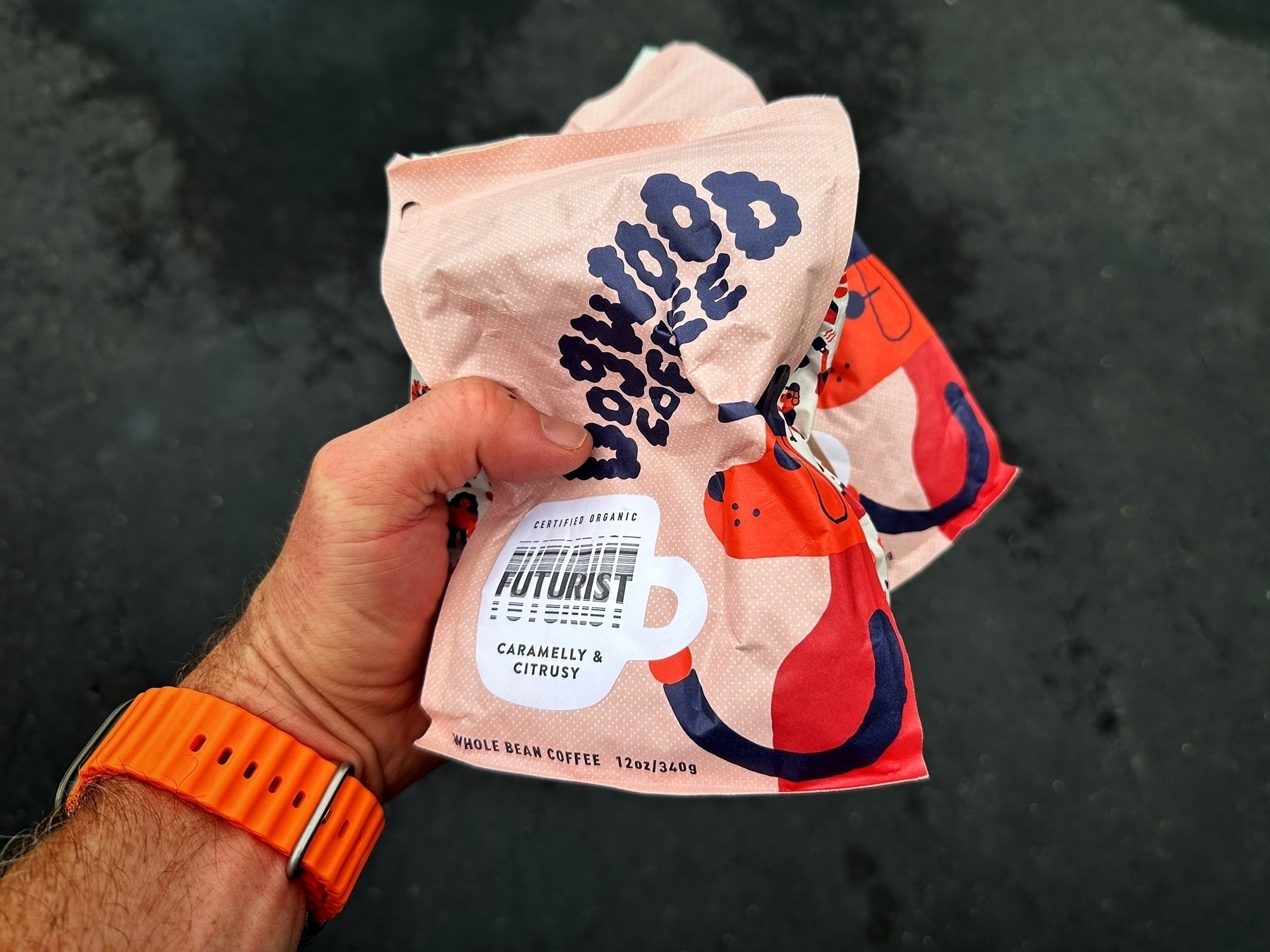 A hand grips two colorful bags of “Outwood Coffee,” featuring “FUTURIST - Caramelly & Citrusy - Whole Bean Coffee, 12oz/340g,” set against a dark, blurred outdoor background.