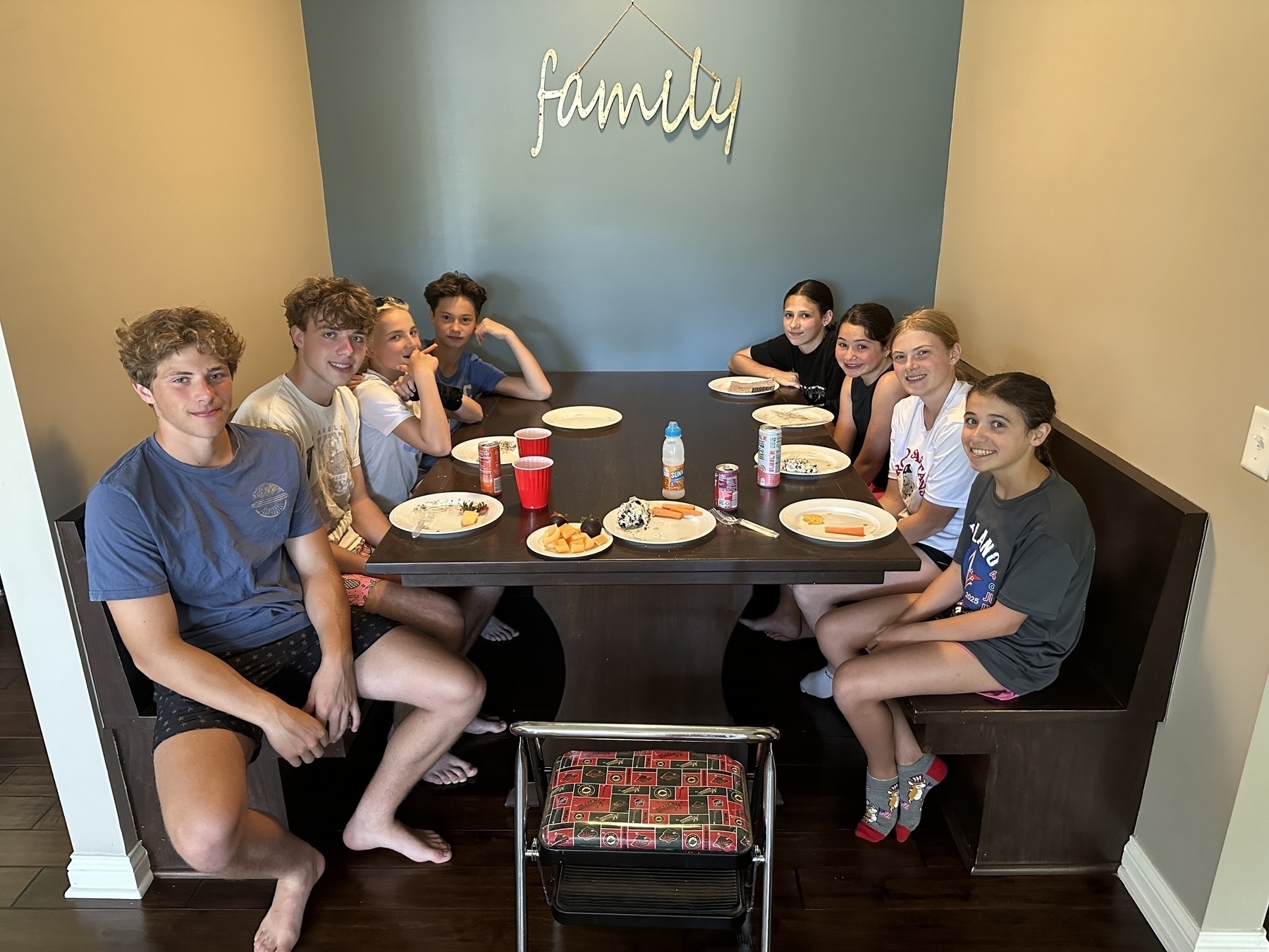 A group of eight people seated around a dining table, enjoying a meal. The wall in the background features a decorative “family” sign. The table has plates with food and various drinks. The room has a cozy and welcoming atmosphere.