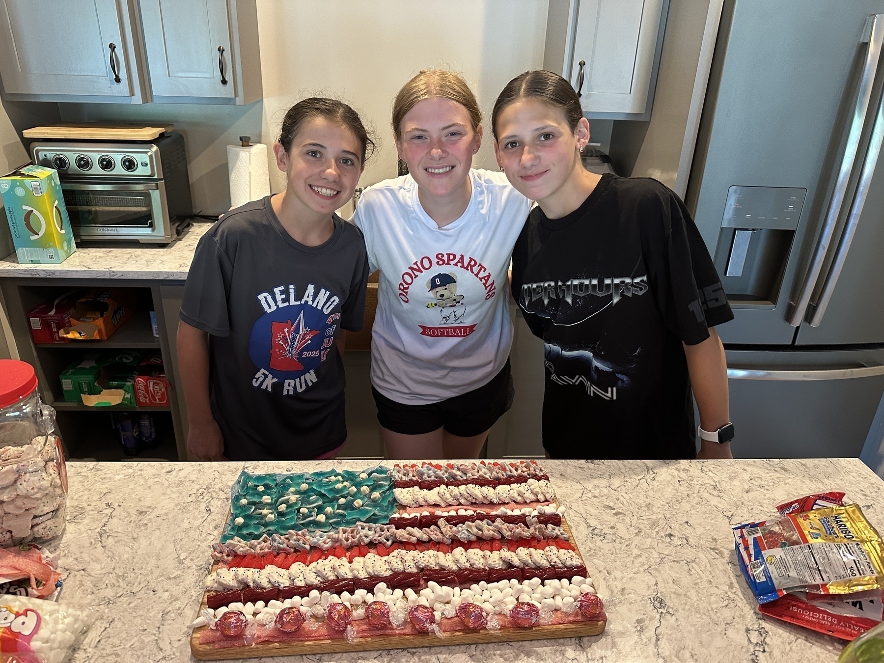 Three smiling individuals stand behind a kitchen counter, displaying a dessert decorated like an American flag using various candies.