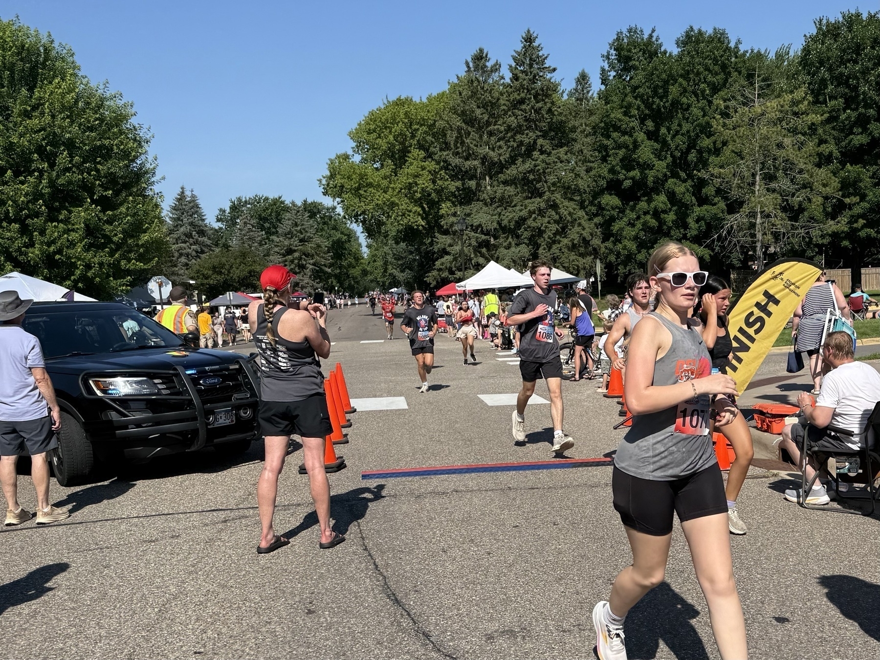 Runners nearing the finish line of a race on a sunny day, with spectators and event staff watching. A finish banner and orange cones are visible, along with a parked police car on the side.