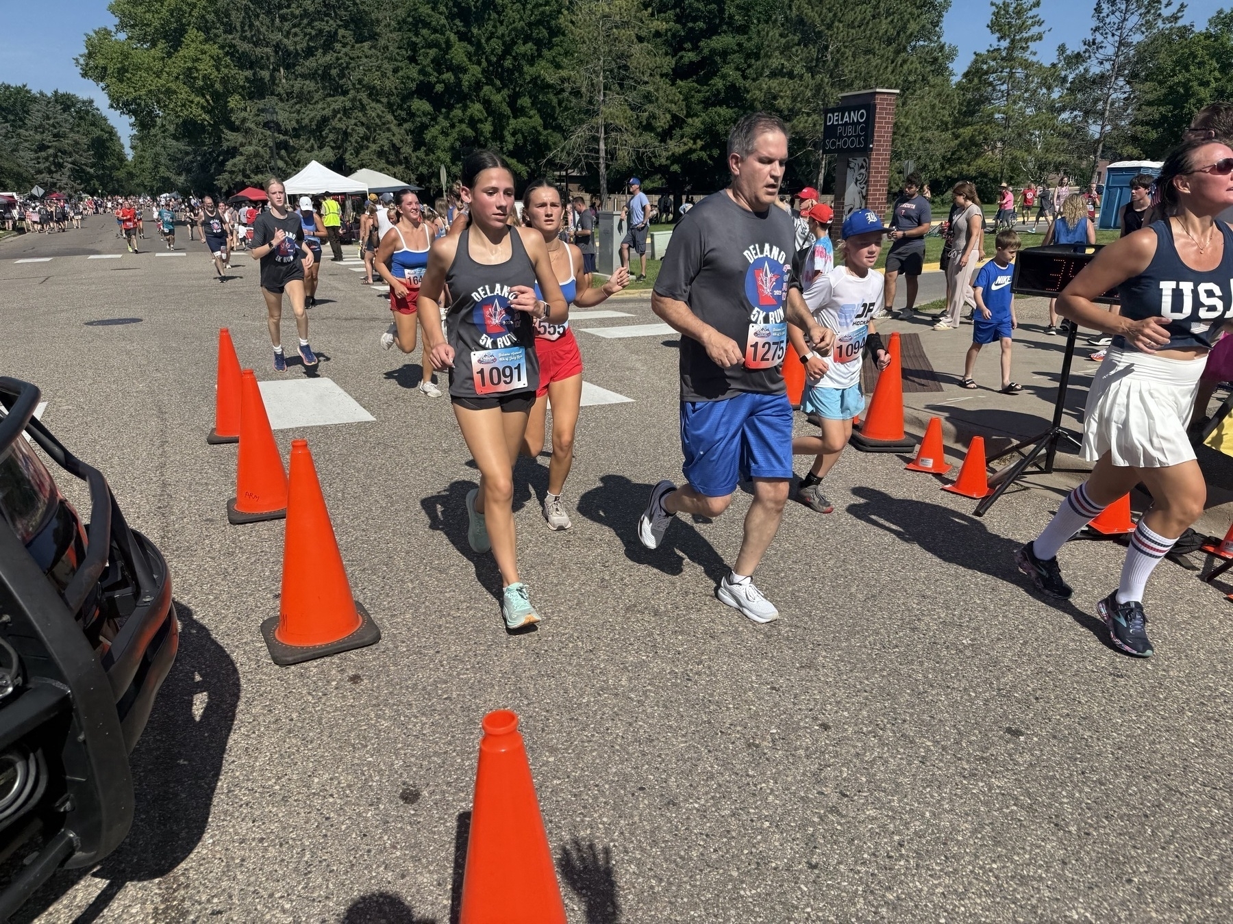Runners participating in a road race, surrounded by orange cones, on a sunny day. The scene is lively with people wearing various sports attire, some displaying race numbers. Tall trees and a school sign are visible in the background.