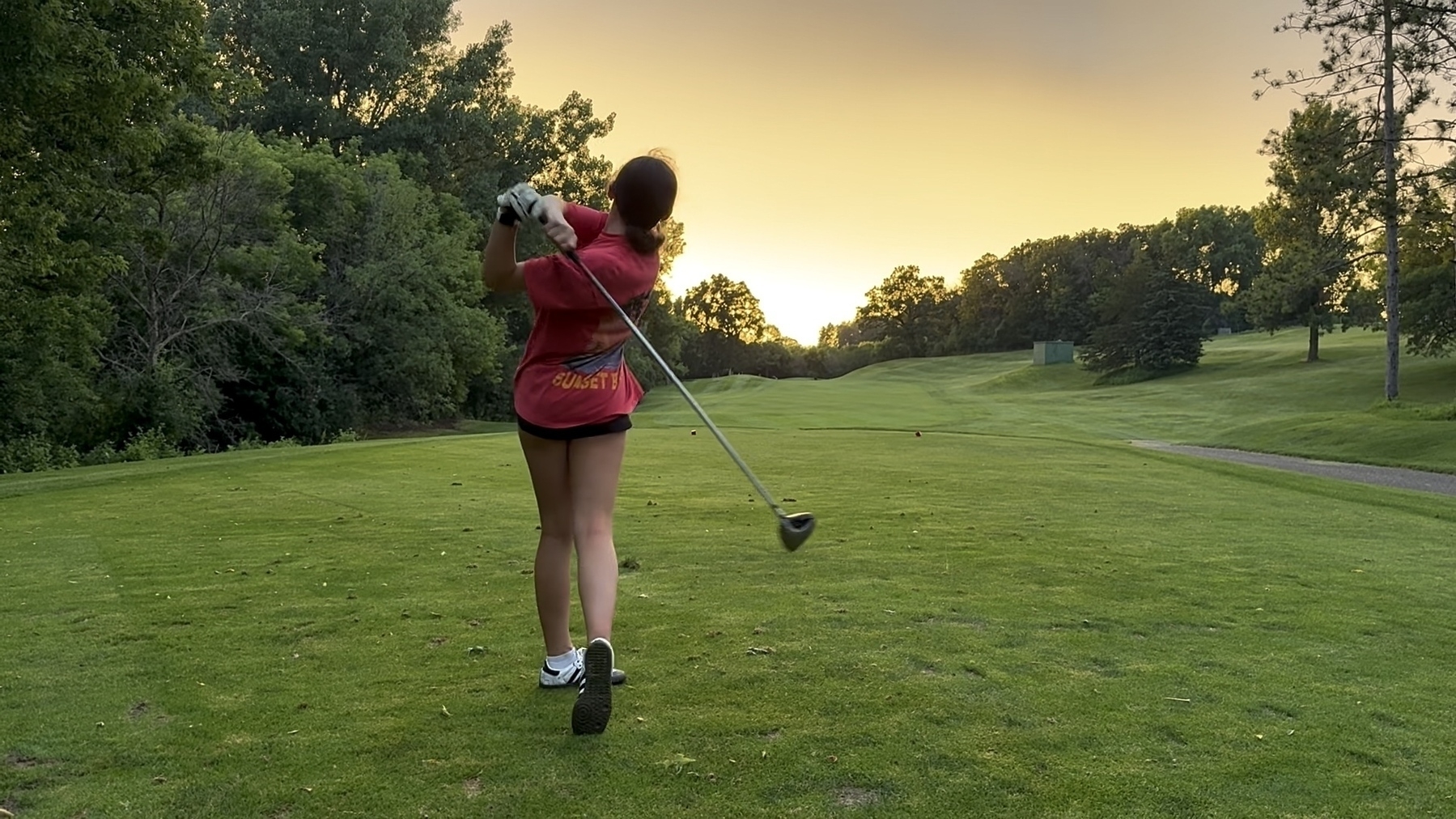 A person in a red shirt swings a golf club on a green fairway during a golden sunset. Trees line the horizon under a clear sky.