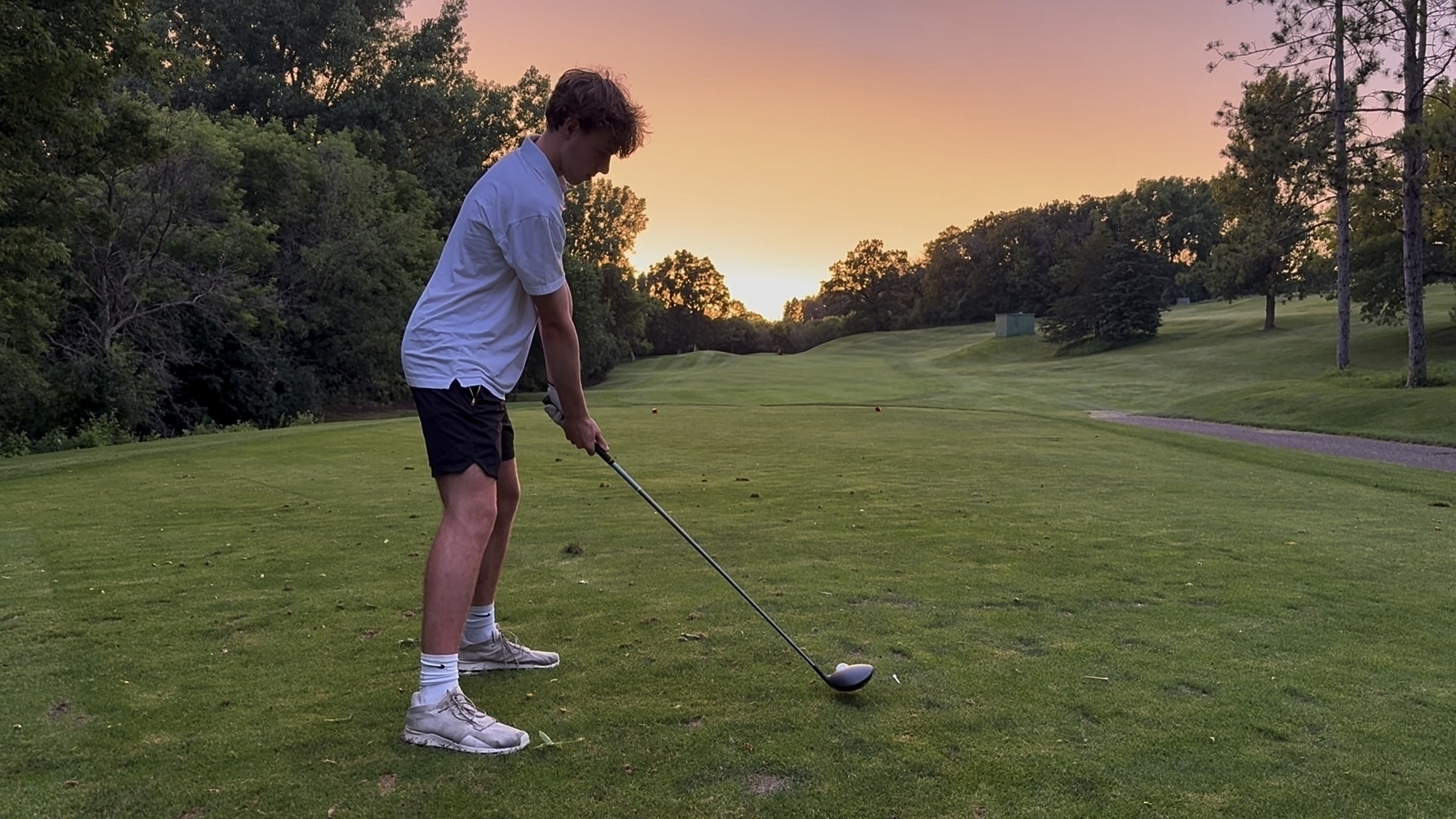 A person in a white shirt and black shorts prepares to swing a golf club on a lush green course at sunset, with trees in the background and a clear path ahead.