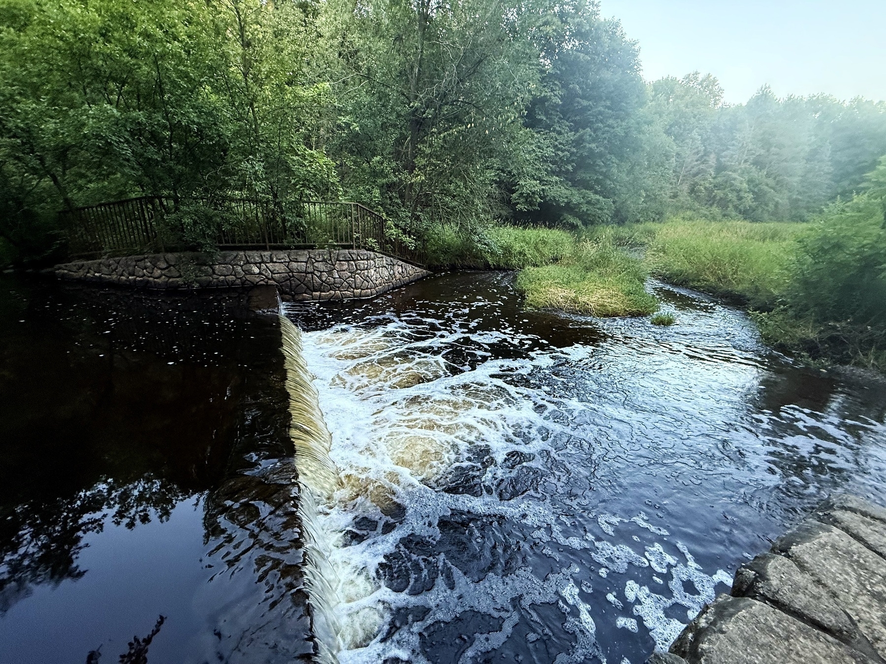 A small waterfall flows over a stone barrier into a calm river, surrounded by lush green trees and vegetation. A stone path with a fence is visible to the left, curving along the riverbank.
