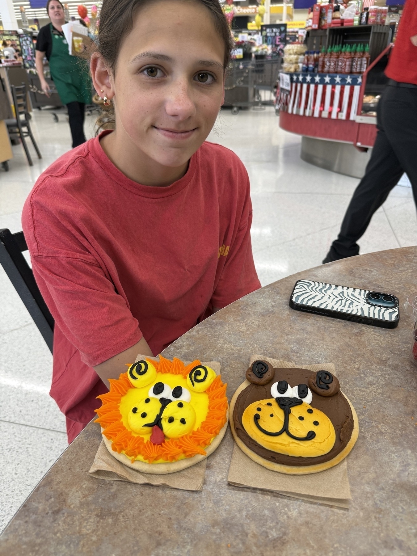 A person wearing a red shirt sits at a table in a store, with two decorated cookies in front of them. One cookie is decorated to look like a lion with orange and yellow icing, and the other like a bear with brown and yellow icing.