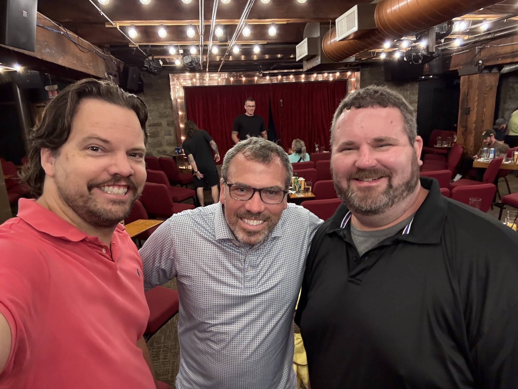 Three people smiling in a cozy venue with red curtains and rows of seating. Ceiling lights create a warm atmosphere.