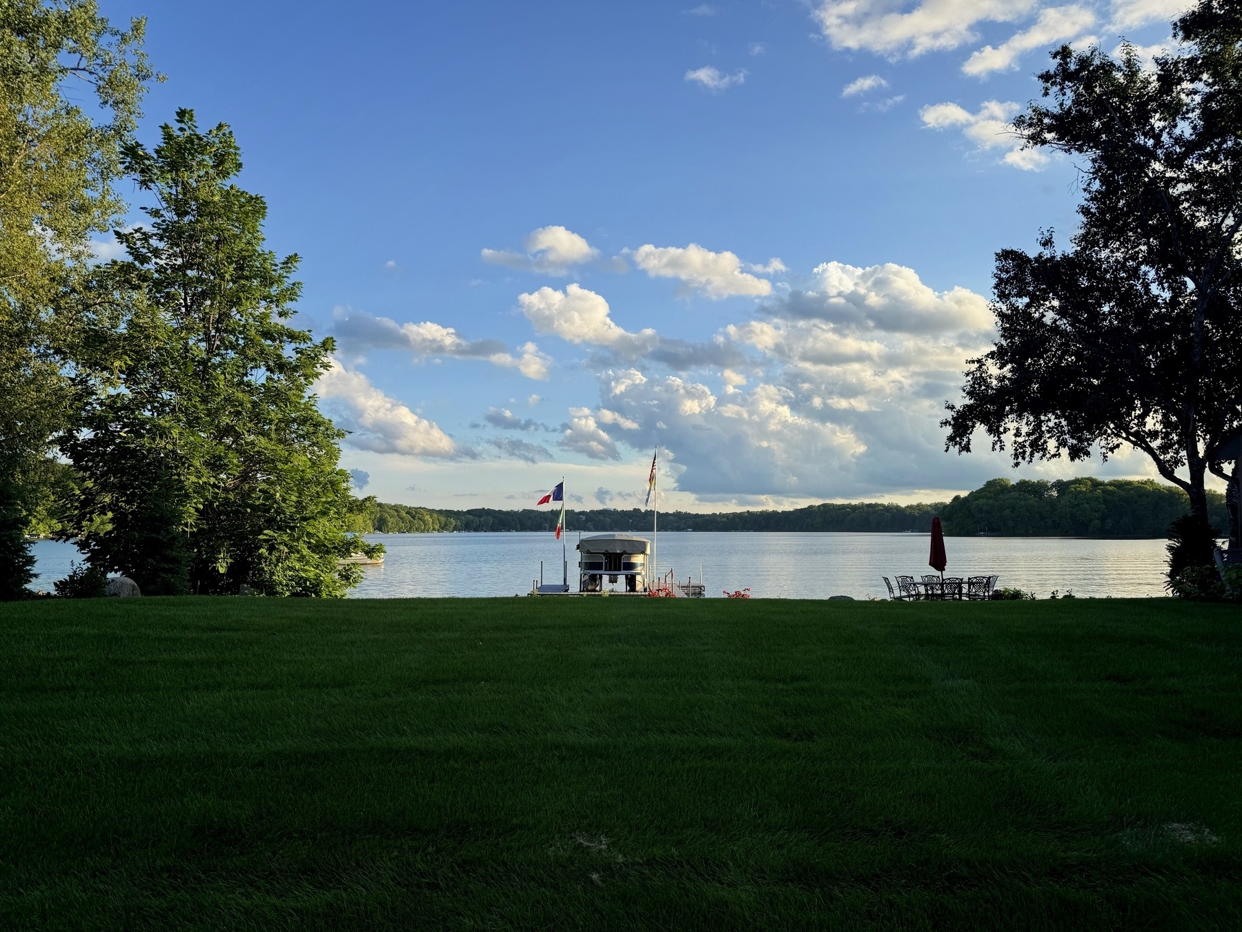 A serene lakeside scene with a well-manicured grassy lawn in the foreground. In the distance, a dock with flags stands at the water’s edge, surrounded by calm waters and bordered by lush green trees. Fluffy clouds are scattered across the blue sky.