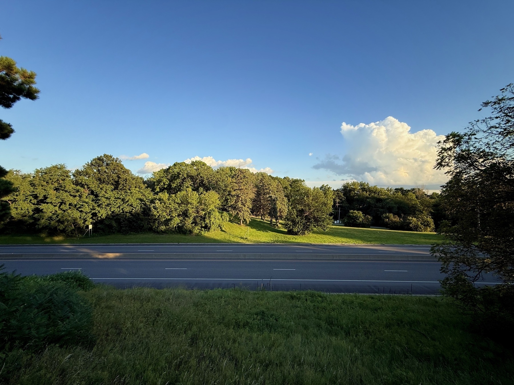 A multi-lane road runs through the foreground, bordered by a grassy area. Beyond the road, a lush, green landscape features dense trees under a clear blue sky with a few clouds.