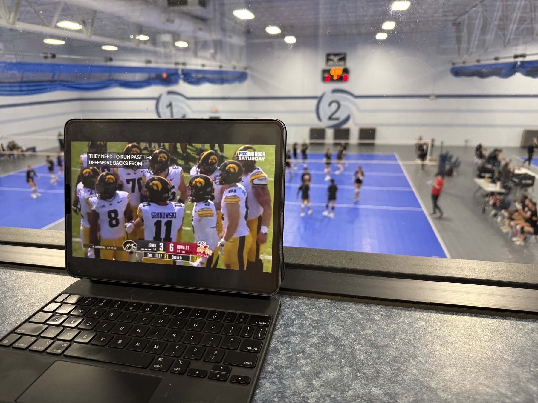 A tablet displaying a football game is placed on a countertop inside a sports facility. In the background, there are volleyball courts with athletes playing.