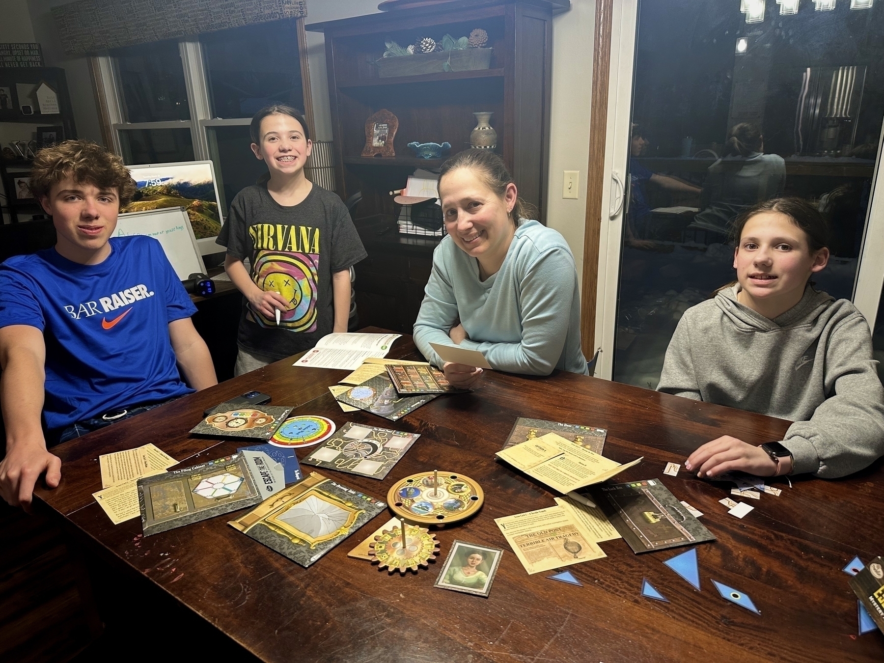 A group of four people sitting around a wooden table playing a board game. The table is covered with game components, including cards and tokens. A shelf with decorations is visible in the background, and a large window reflects indoor lighting.
